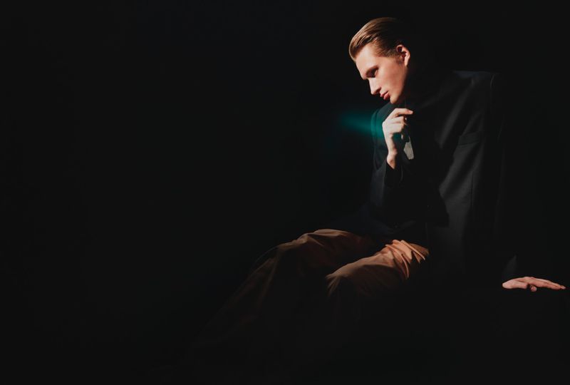 A man in a dark tailored blazer and tan trousers seated against a dark background in dramatic low-key lighting.
