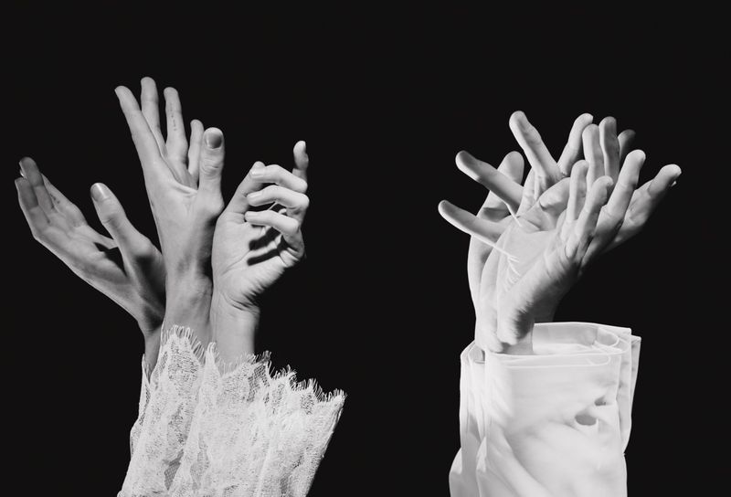 Black and white photograph of multiple hands emerging from white lace and cotton shirt cuffs against a black background.