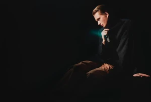 A man in a dark tailored blazer and tan trousers seated against a dark background in dramatic low-key lighting.