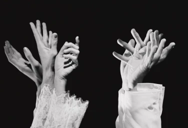 Black and white photograph of multiple hands emerging from white lace and cotton shirt cuffs against a black background.