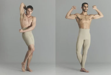 Two shirtless male models posing in beige high-waisted tights against a neutral grey studio background.
