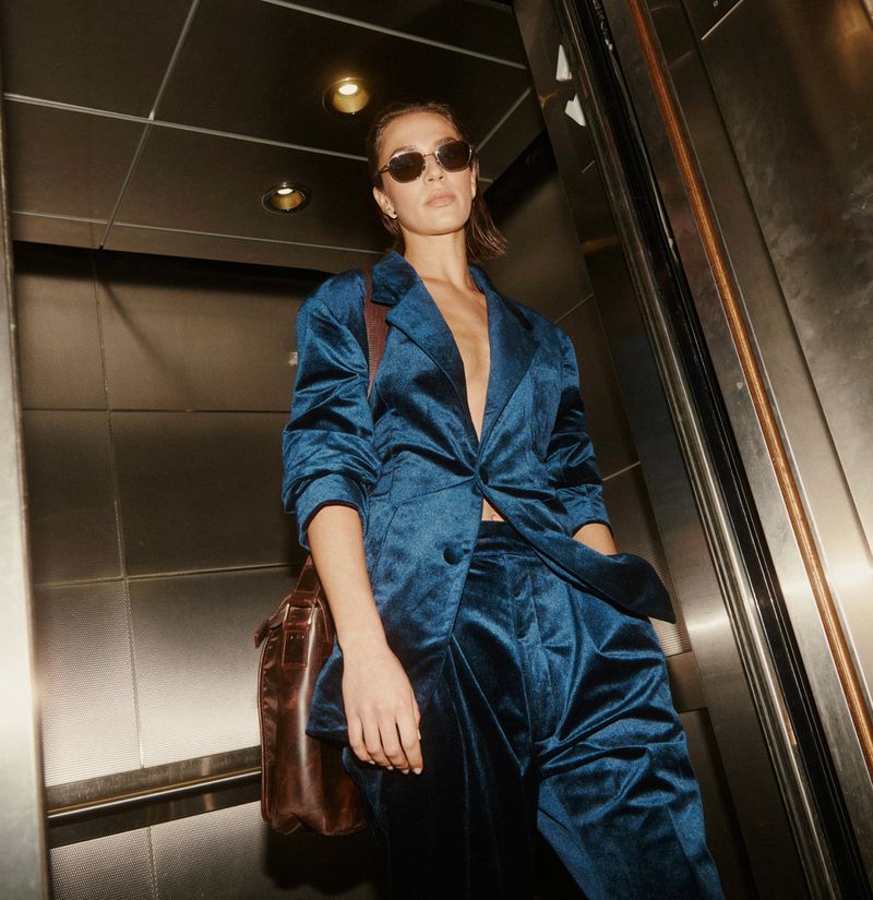 Woman wearing a petrol blue velvet suit and dark sunglasses carrying a brown leather shoulder bag inside a metal elevator.