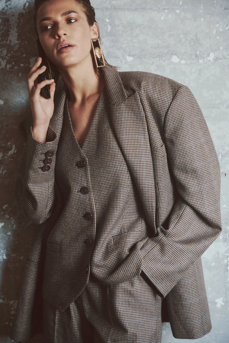 Model wearing an oversized brown houndstooth three-piece suit and statement earrings against a textured concrete wall.
