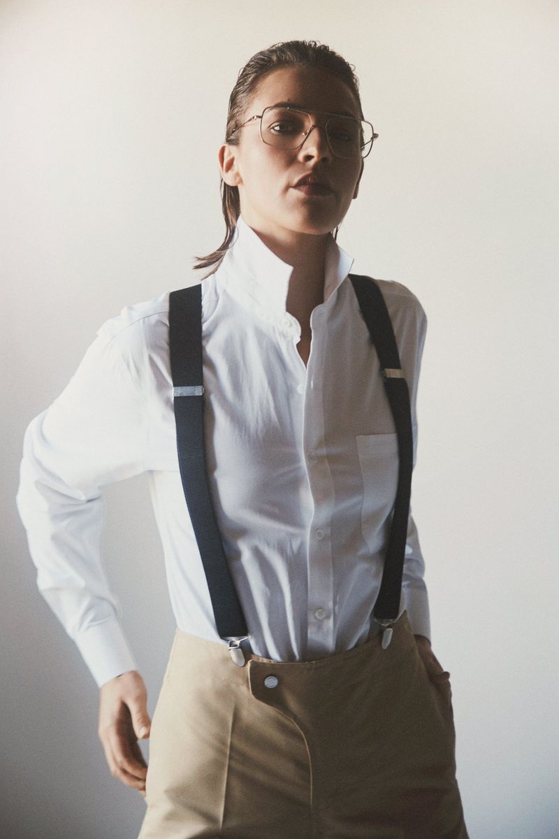 Woman in wireframe glasses wearing a white shirt with black suspenders and beige trousers against a neutral background.