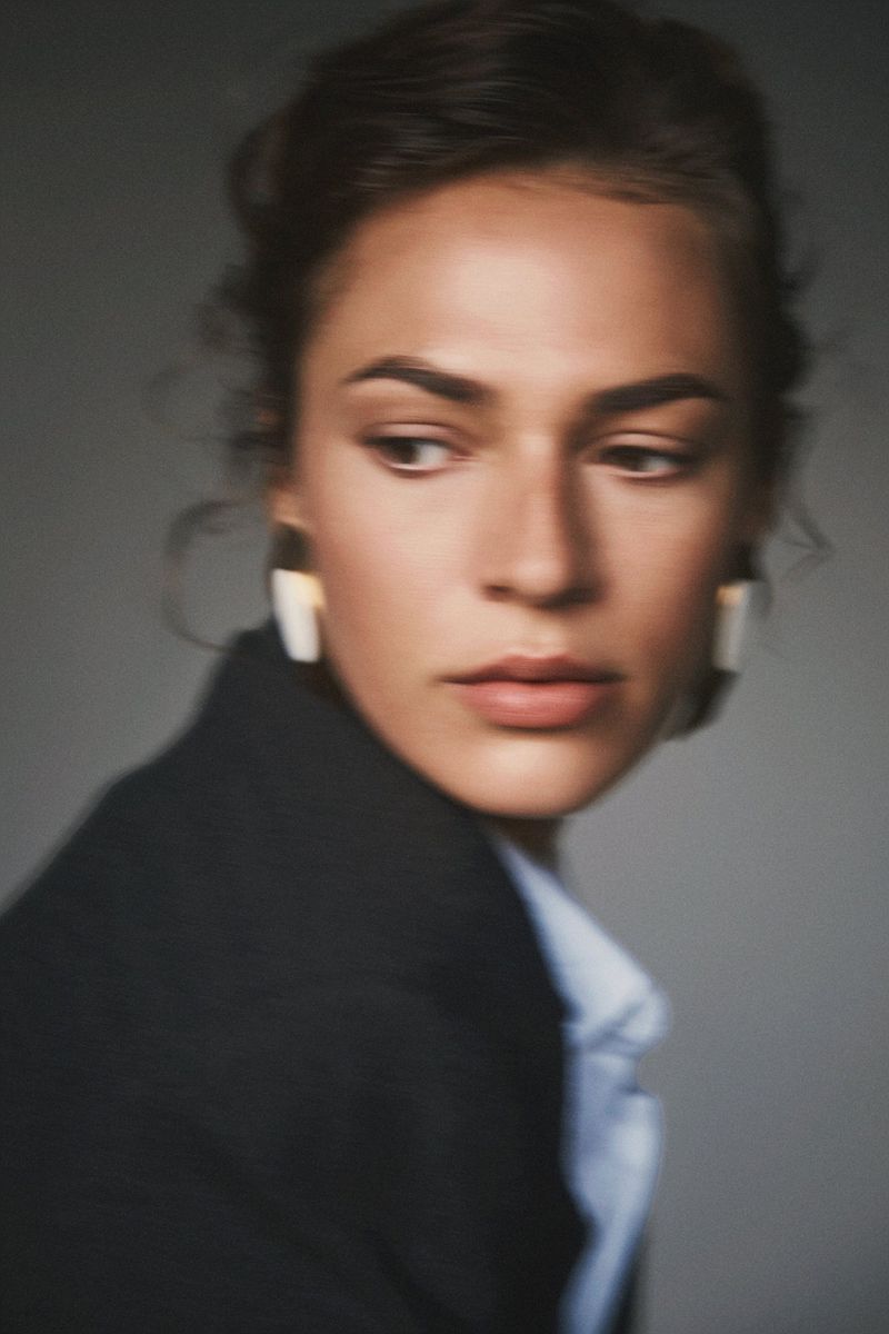 Woman looking over her shoulder wearing a black tailored blazer, light blue collared shirt, and bold statement earrings against a grey backdrop.