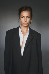 Woman wearing an oversized black blazer and open white collared shirt against a grey studio backdrop.