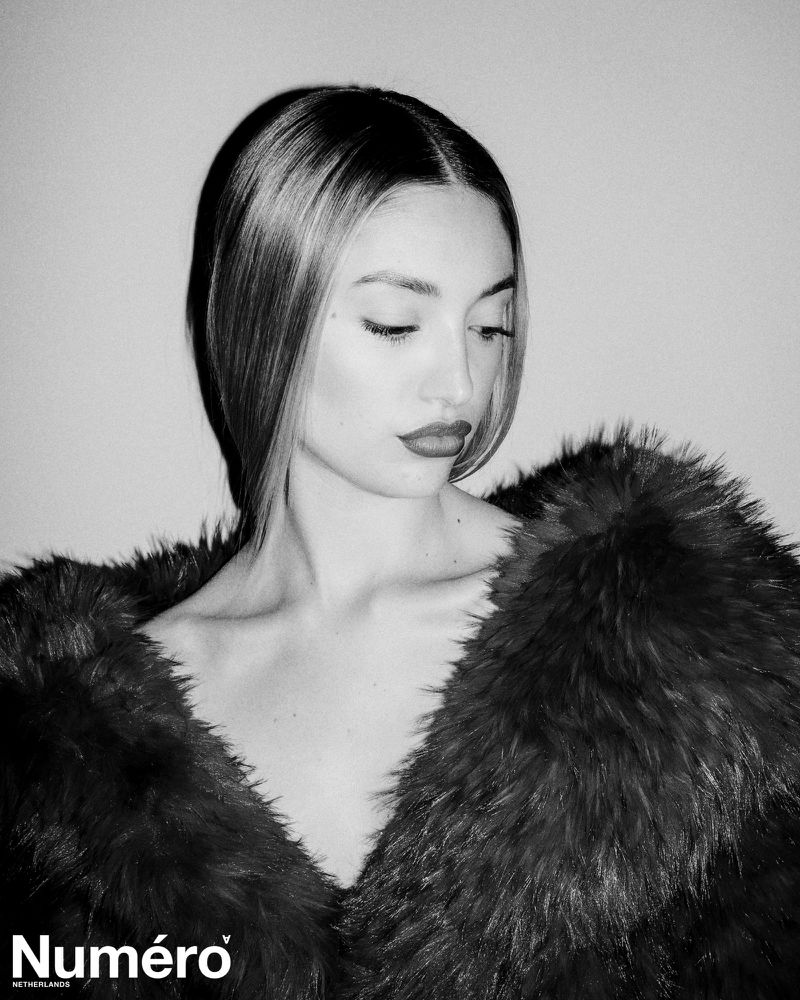 Black and white portrait of Bella Podaras looking downward while wearing a voluminous dark fur collar.
