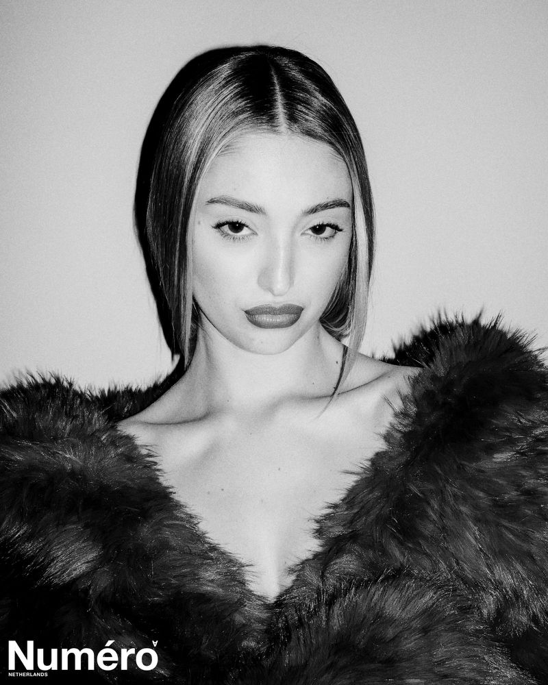 Black and white portrait of Bella Podaras wearing a textured fur coat against a plain backdrop with direct flash lighting.