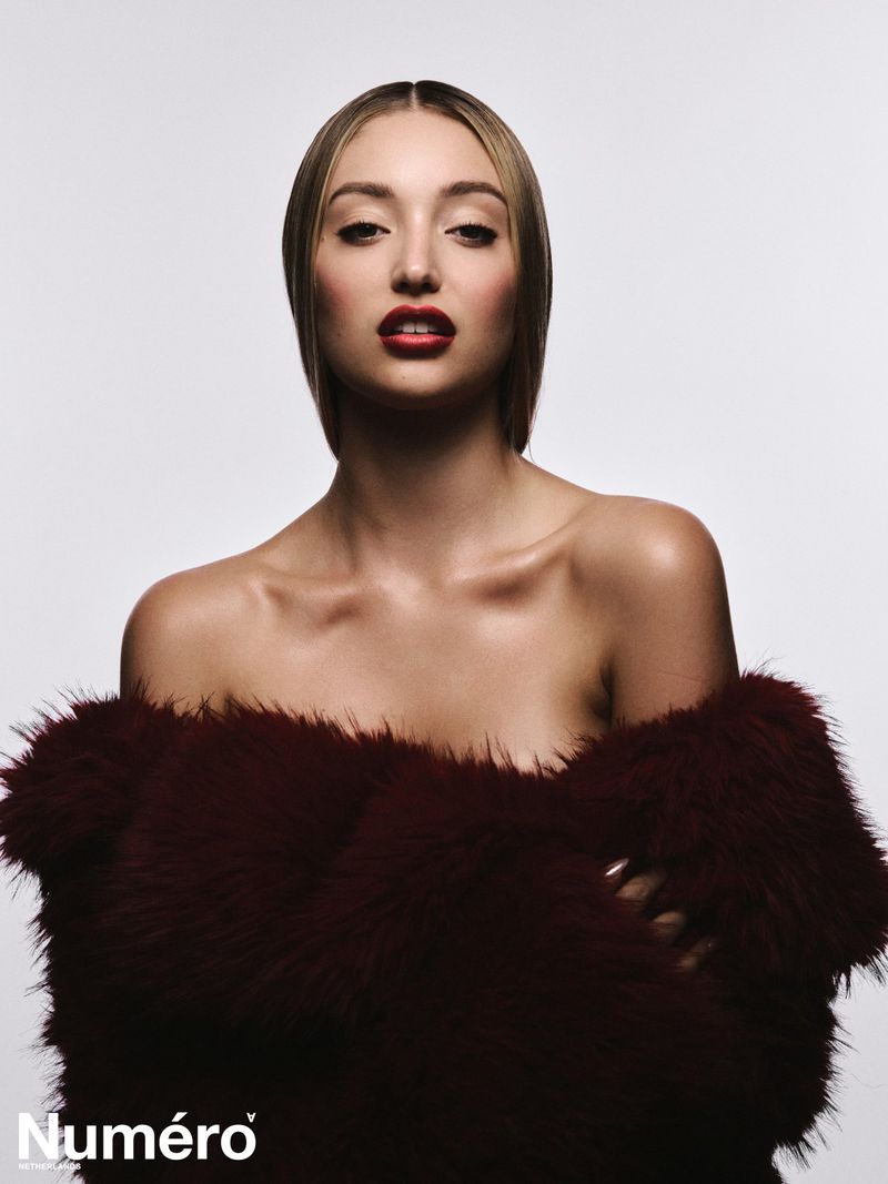 Bella Podaras posing with bare shoulders wrapped in a dark burgundy faux fur coat against a white studio background.