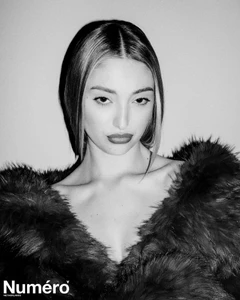 Black and white portrait of Bella Podaras wearing a textured fur coat against a plain backdrop with direct flash lighting.