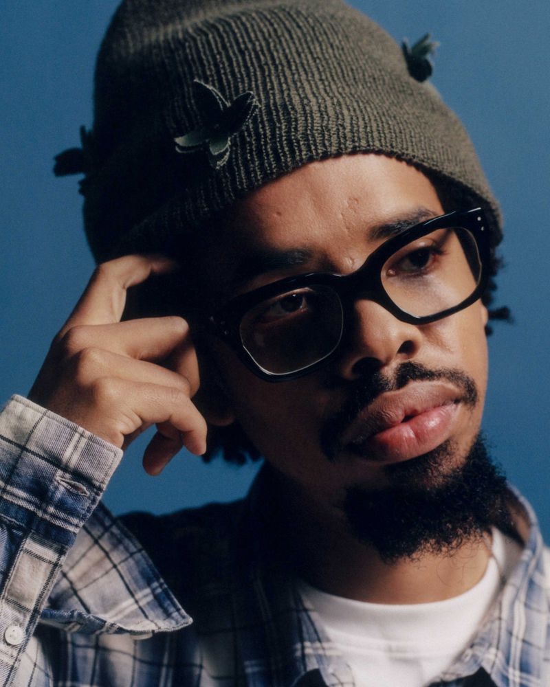 Earl Sweatshirt wearing a green beanie with butterfly details, black glasses, and a plaid shirt against a blue background.