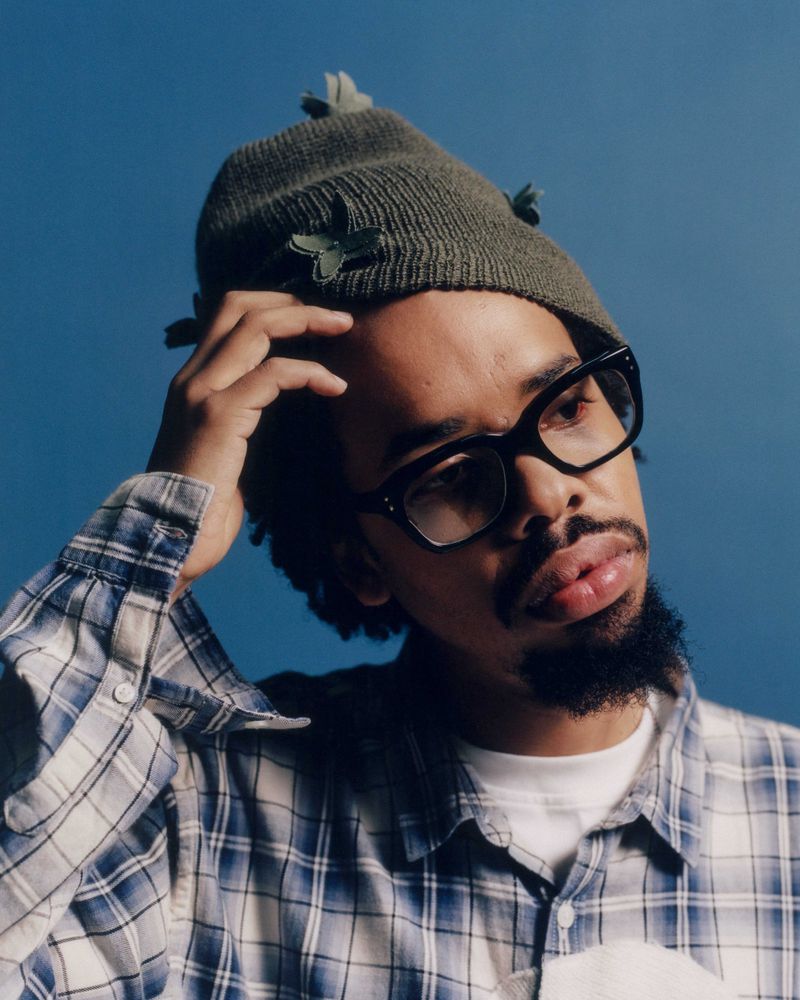 Earl Sweatshirt wearing black glasses, an olive knit beanie, and a blue plaid shirt against a blue backdrop.