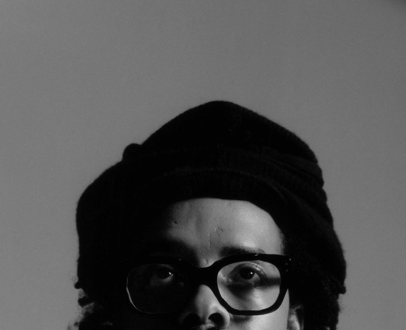 Portrait close-up of Earl Sweatshirt wearing thick-framed glasses and a beanie against a plain neutral background.