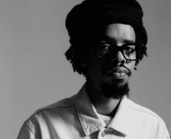 Black and white portrait of Earl Sweatshirt wearing dark glasses, a knit hat, and a light collared jacket.