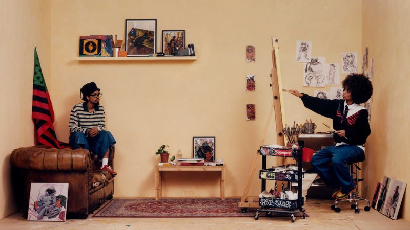 Earl Sweatshirt seated on a leather armchair while artist Bianca Simone Scott paints at an easel in a warm studio space.