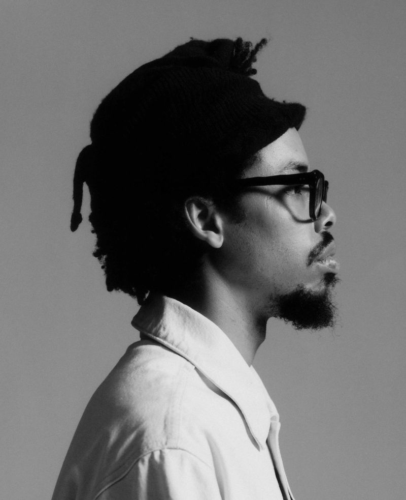 Black and white profile portrait of Earl Sweatshirt wearing glasses and a light collared shirt against a grey background.