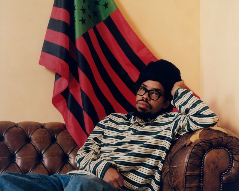 Earl Sweatshirt reclining on a worn brown leather sofa wearing a striped long-sleeve shirt and black beanie under a Pan-African flag.