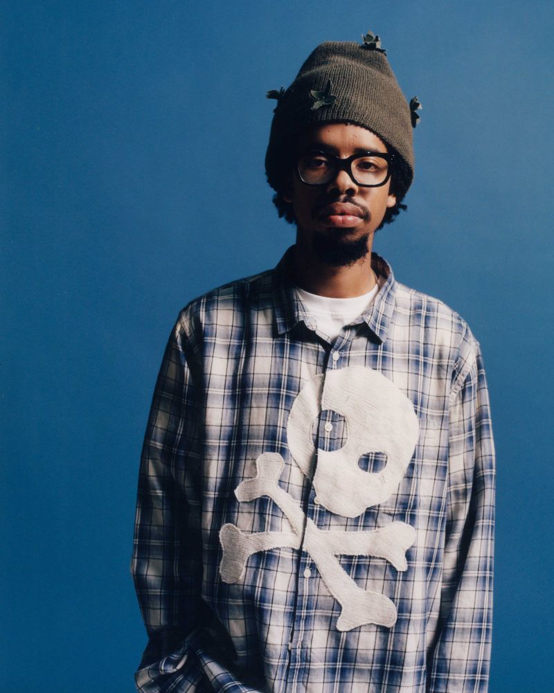 Earl Sweatshirt wearing a beanie, glasses, and a plaid shirt with a skull patch against a solid blue background.