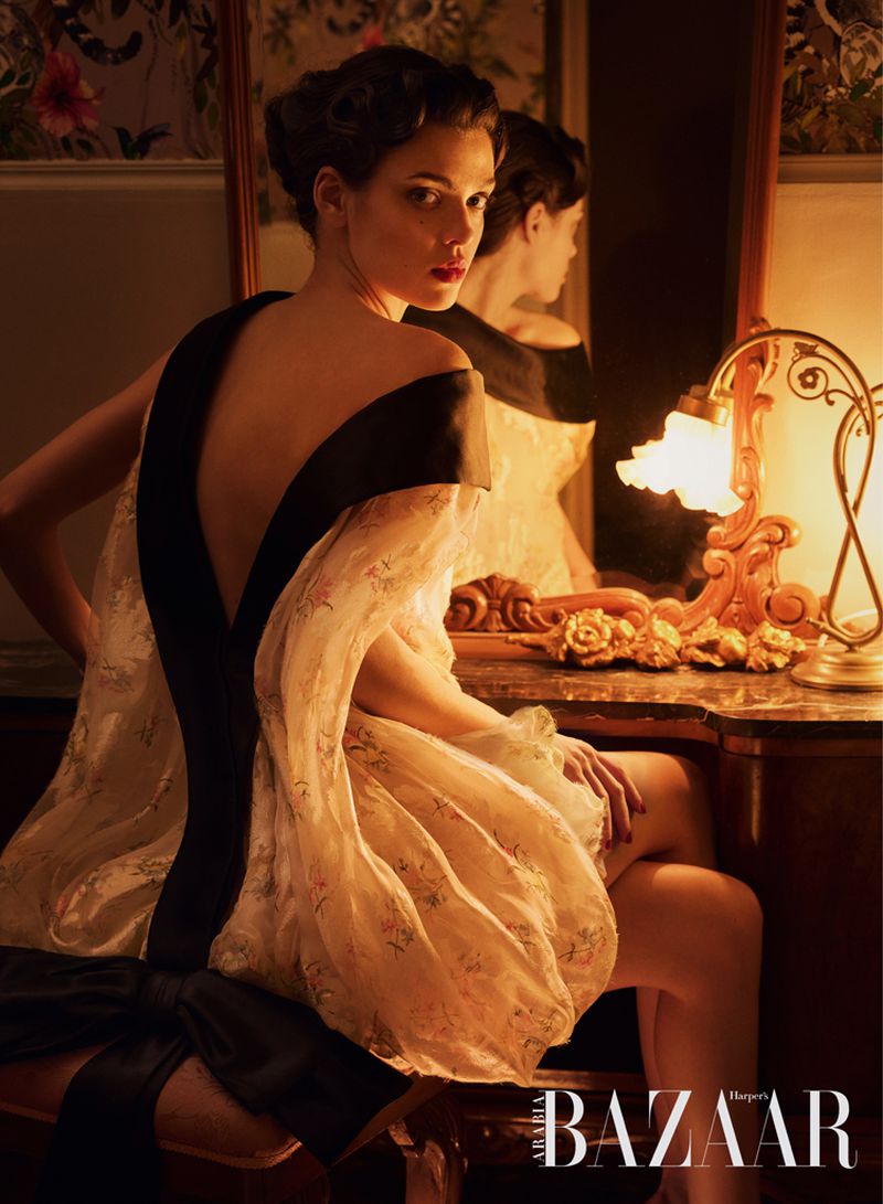 Woman in backless floral dress with black trim sitting at an antique vanity mirror illuminated by warm lamp light.