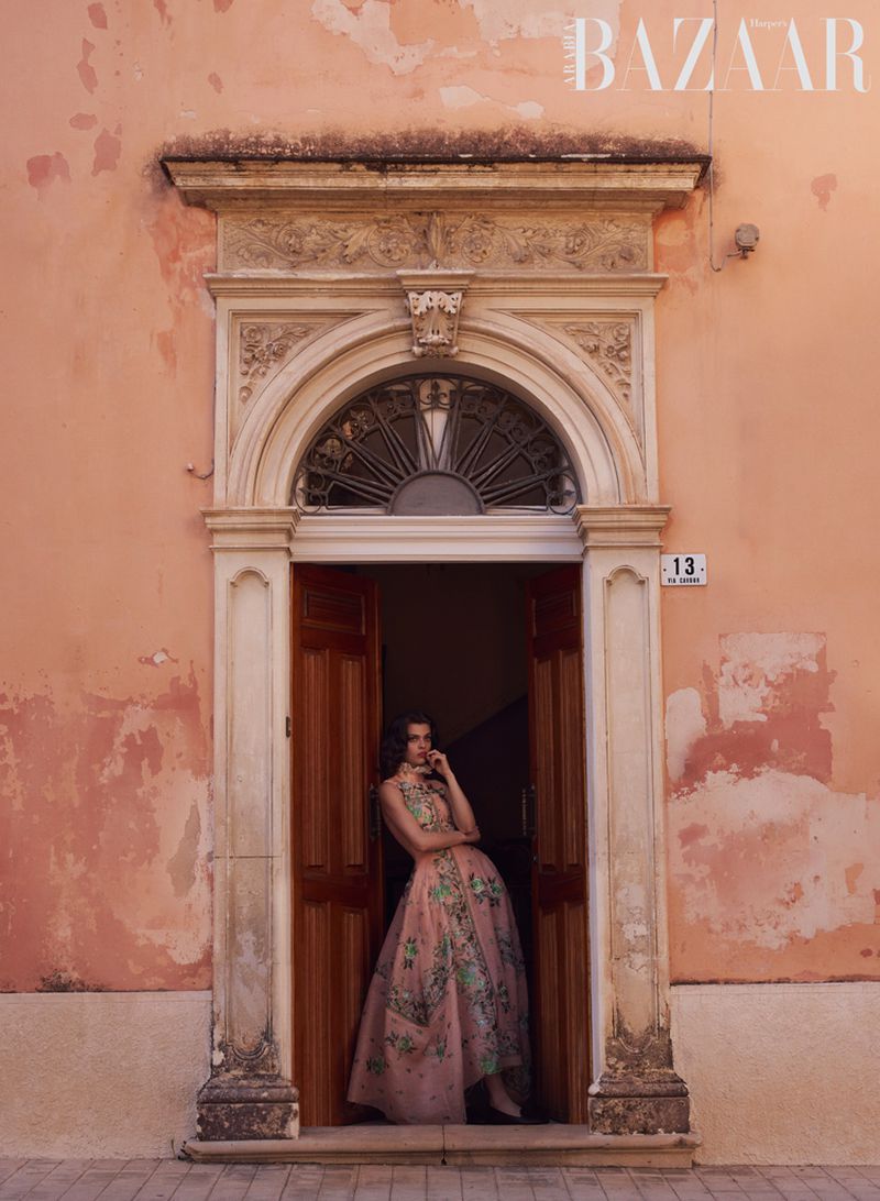 Model wearing a floral embroidered pink evening gown standing in an ornate classical arched doorway of a peach plaster building.