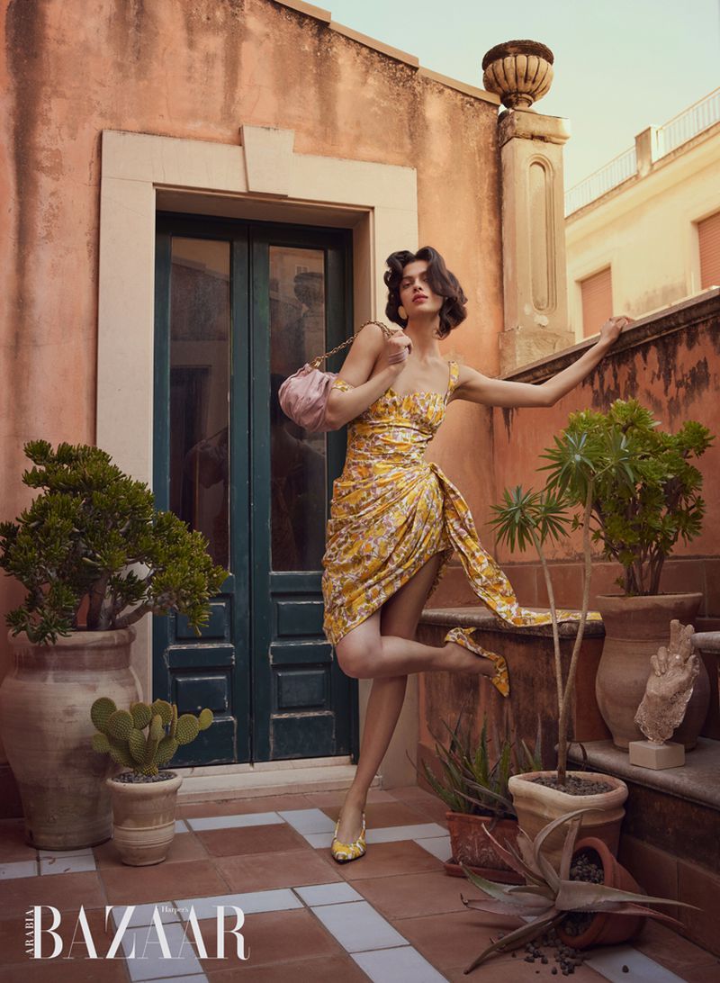 Model posing in a yellow floral draped dress and matching heels holding a pink handbag on a sunlit terracotta terrace.
