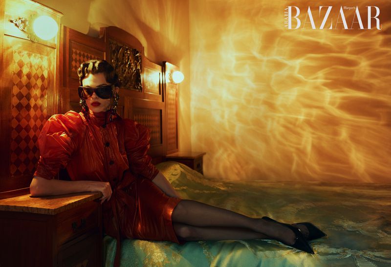 Model in a shiny red puff-sleeve dress, sunglasses, and black heels reclining on a bed under warm amber lighting.