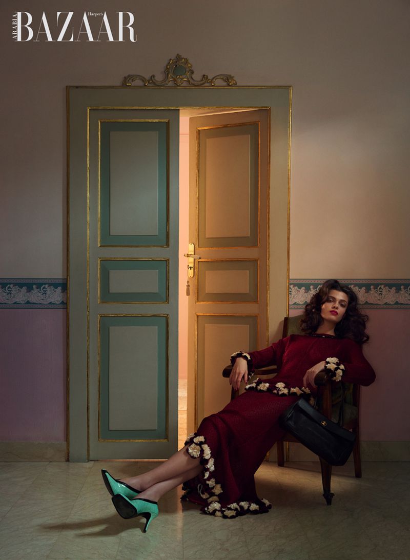 Model in a burgundy dress with floral trim and mint green heels seated on an armchair beside ornamental double doors.
