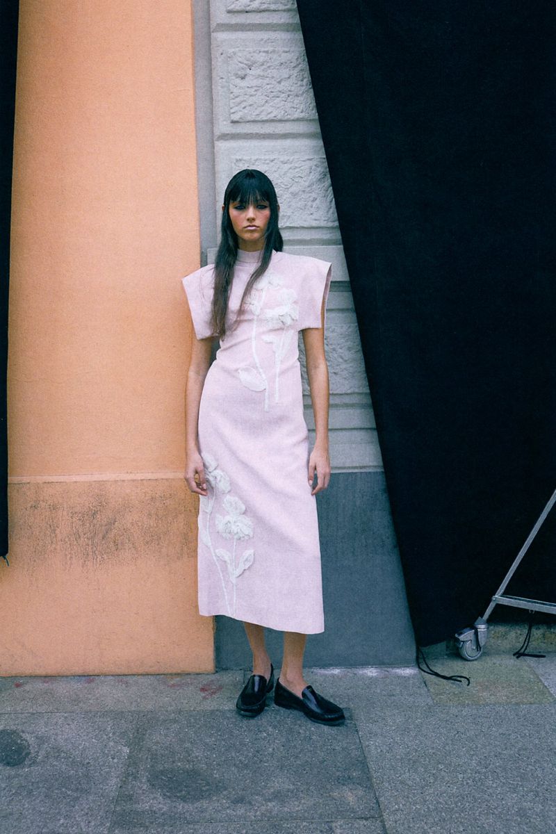 A fair-skinned adult fashion model with bangs poses in a pale dress with floral appliqué on a city sidewalk next to a peach and black wall.