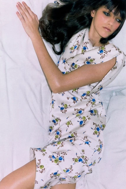 Brunette woman lying on a white sheet wearing a white floral dress with blue and green flower patterns, posing for a fashion shot.