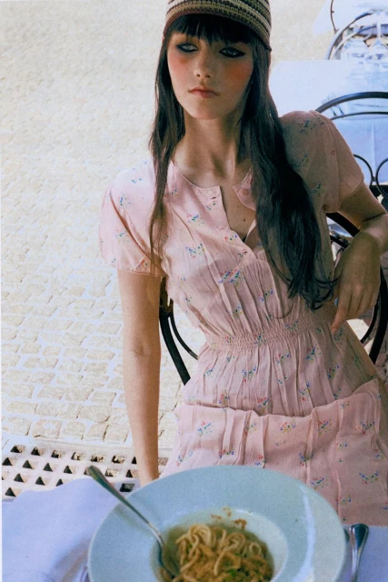 A fair-skinned woman in a light pink floral dress and striped hat sits at an outdoor table with a plate of spaghetti.
