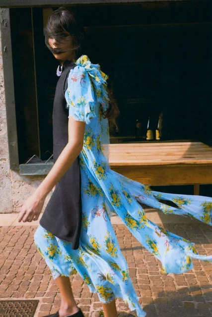 A woman models a blue floral chiffon dress with a black vest walking on a brick path outside on a sunny day.