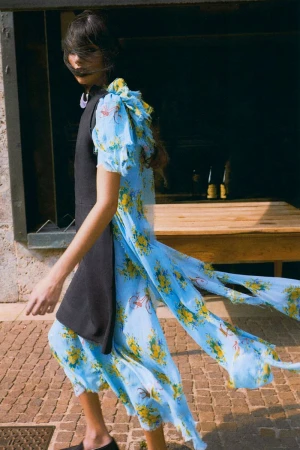 A woman models a blue floral chiffon dress with a black vest walking on a brick path outside on a sunny day.
