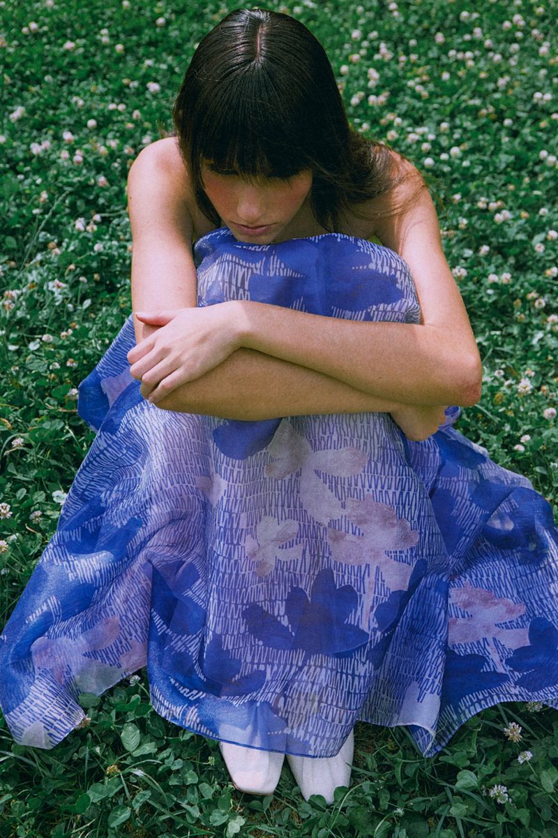 Young adult female in a blue floral dress sitting in a green clover field, hugging her knees in an outdoor setting, eye level shot.
