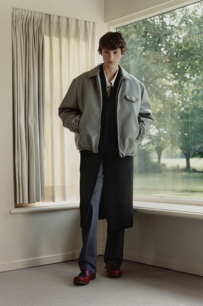 Model Gaspard Sandor standing indoors by a window wearing an oversized grey jacket over a long black layer and tailored trousers.
