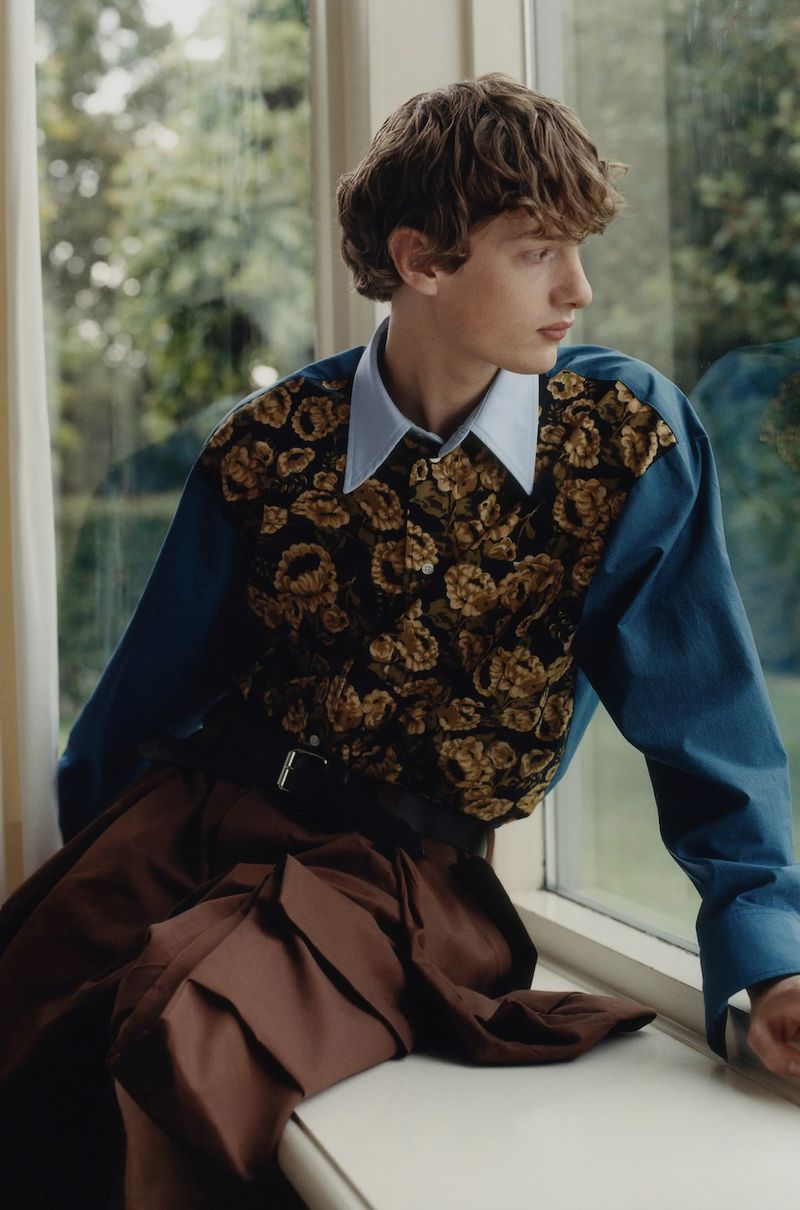 A young male model seated on a windowsill wearing a patterned floral vest over a light blue shirt and brown trousers.