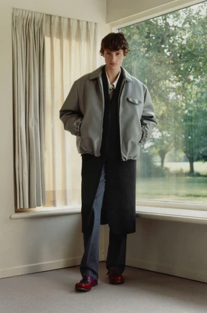 Model Gaspard Sandor standing indoors by a window wearing an oversized grey jacket over a long black layer and tailored trousers.