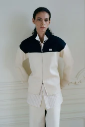 Model Leon Dame wearing a black and cream zip-up jacket and striped shirt for Wales Bonner against a white paneled wall.