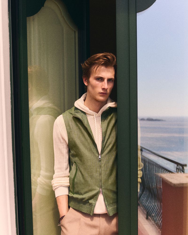 Male model in a green vest and white hoodie standing in a doorway overlooking the sea for Brett Johnson Spring Summer 2027.