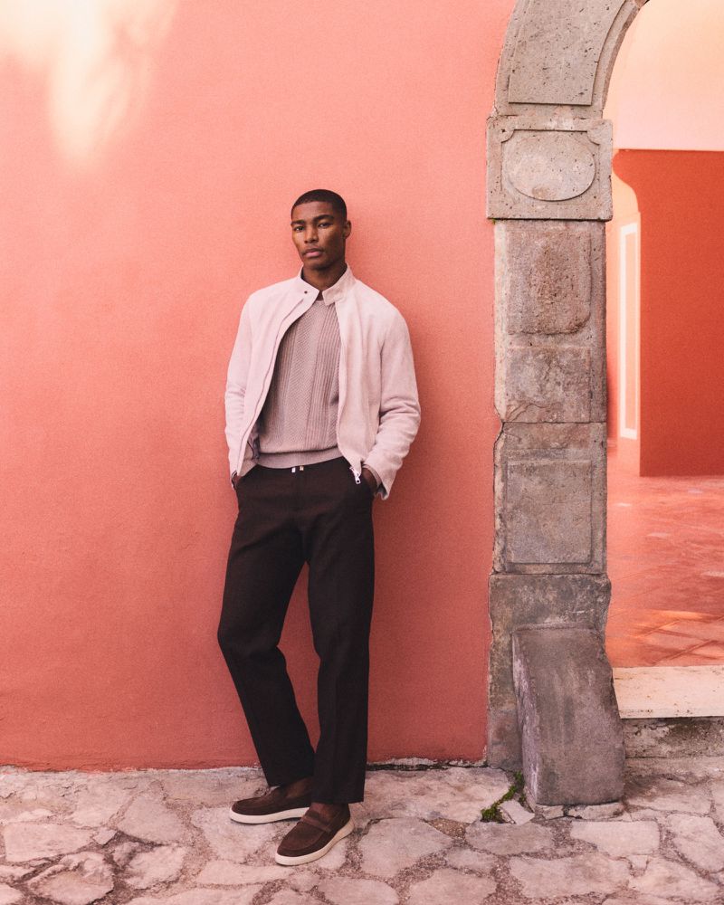 Model Niyo Malik poses in a cream jacket, knit sweater, and dark trousers beside a stone arch and terracotta wall.