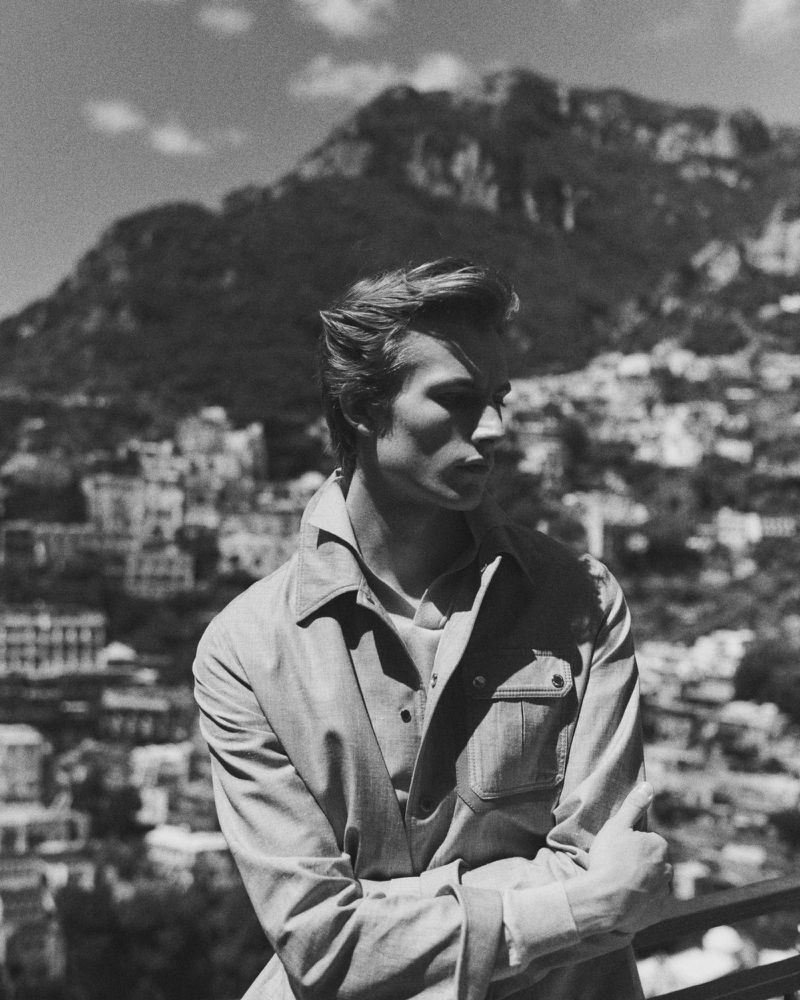 Black and white portrait of male model Oden Delargy wearing a collared utility jacket with a mountain hillside in the background.