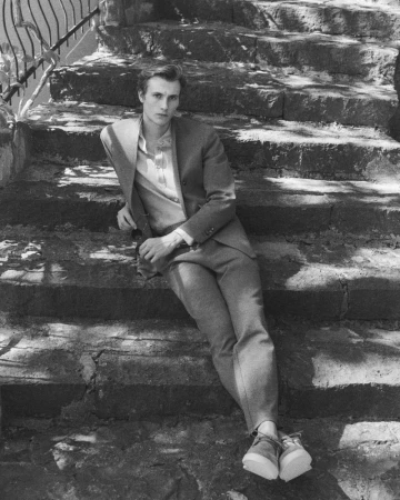 A man in a tailored suit seated on stone outdoor stairs wearing casual shoes for Brett Johnson Spring Summer 2027.