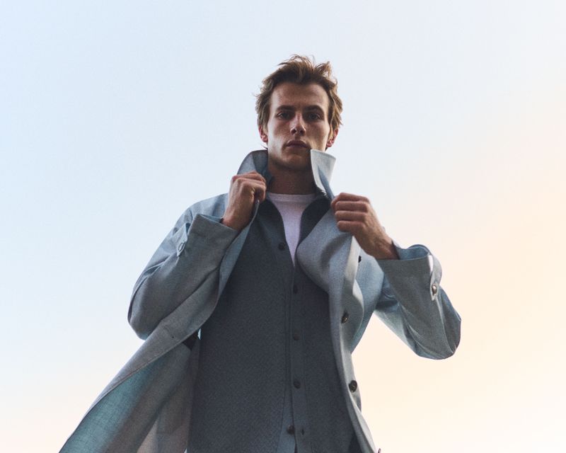 Low-angle shot of a male model wearing a light blue coat and layered shirt against a pale sky.