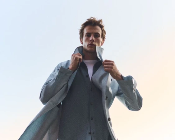Low-angle shot of a male model wearing a light blue coat and layered shirt against a pale sky.