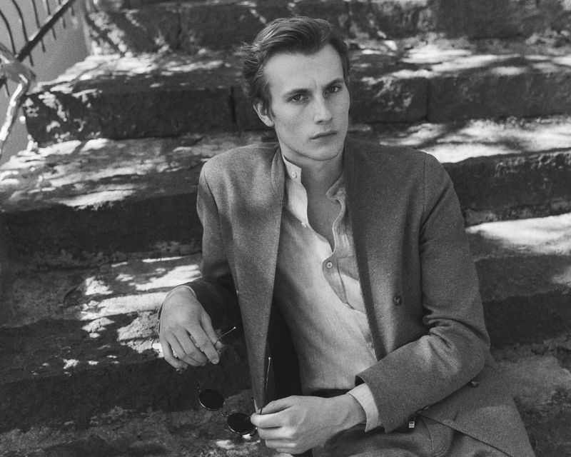 Male model seated on outdoor stone steps wearing a tailored jacket and band-collar shirt while holding round sunglasses.