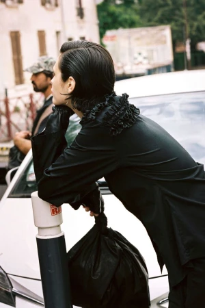 Model Alicia Lindén Guinez wearing a ruffled black shirt while leaning on a street post beside a white car.