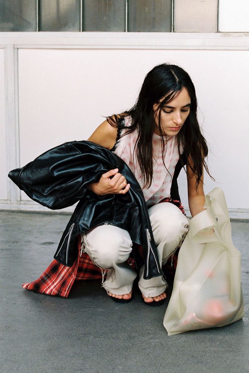Model Alicia Lindén Guinez crouching in distressed white trousers, holding a black leather jacket beside a translucent tote bag.