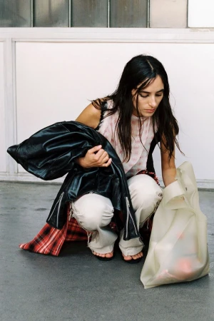 Model Alicia Lindén Guinez crouching in distressed white trousers, holding a black leather jacket beside a translucent tote bag.