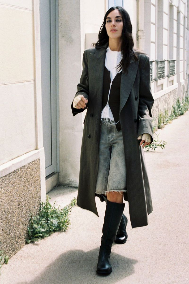 Model in long grey coat, denim shorts, and black boots walking down a city street for British Vogue Our Legacy Spring 2027 menswear project