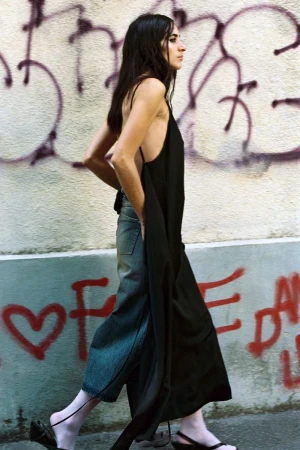 Model Alicia Lindén Guinez walking in profile wearing a black backless tunic over wide-leg denim against a graffiti wall.