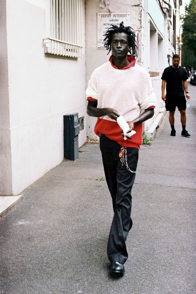 Model walking outdoors along a street wearing a layered cream and red knit sweater paired with black trousers.